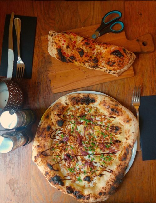 Overhead view of two rustic wood-fired pizzas on a wooden table, one topped with herbs and cured meat, beside plates, cutlery, and scissors.