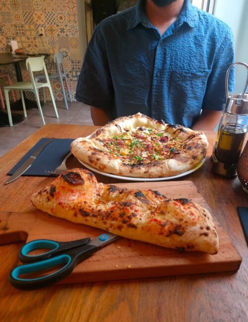 A diner in a blue shirt sits behind two wood-fired pizzas on wooden boards: a round herb-topped pizza and a long, cheesy folded one.