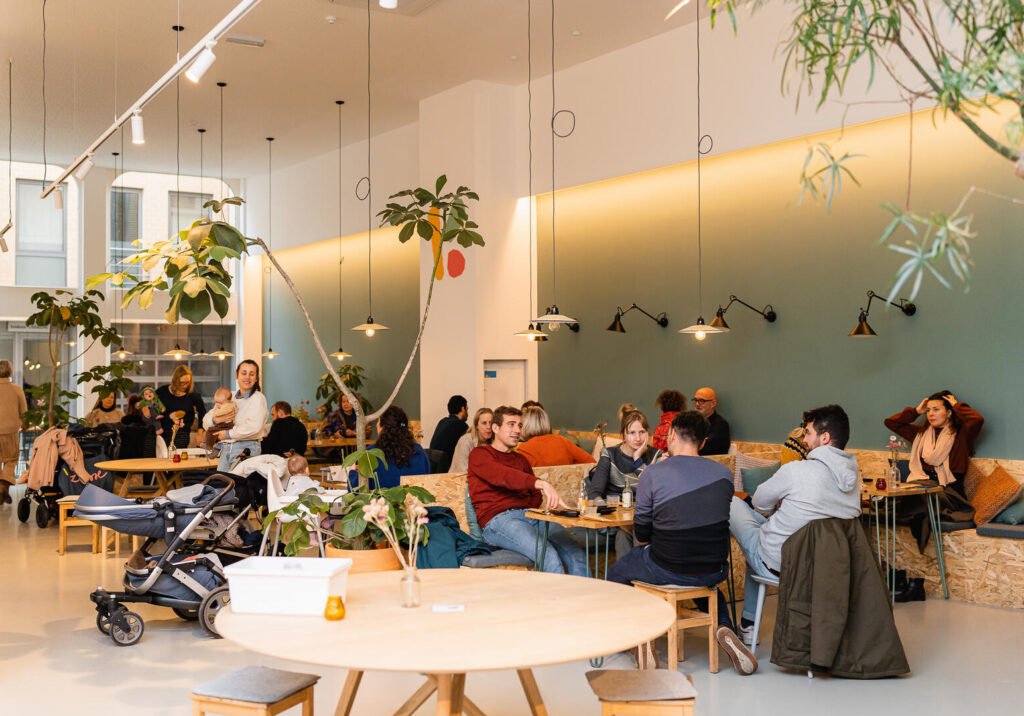 Bustling modern café with diverse groups dining at tables, a baby stroller, lush plants, warm pendant lights, and a cozy atmosphere.