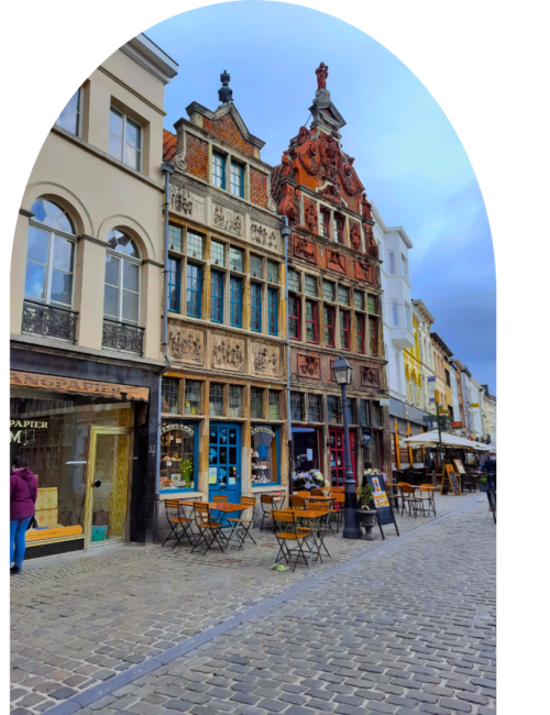 Historic European cobblestone street lined with ornate gabled buildings, shopfronts, and outdoor café seating beneath a bright blue sky.