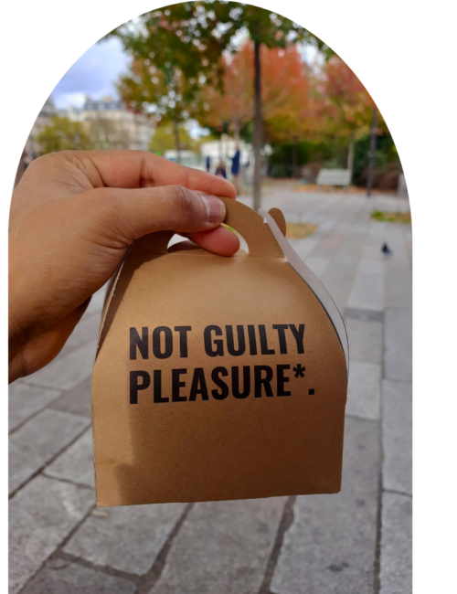 Hand holds a brown takeaway food box labeled “NOT GUILTY PLEASURE*” on a city sidewalk, with autumn trees and pedestrians blurred behind it.