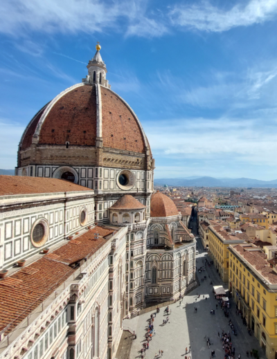 Florence skyline with Brunelleschi’s red-tiled dome and cathedral in foreground, viewed above sunlit streets and historic rooftops under blue sky.