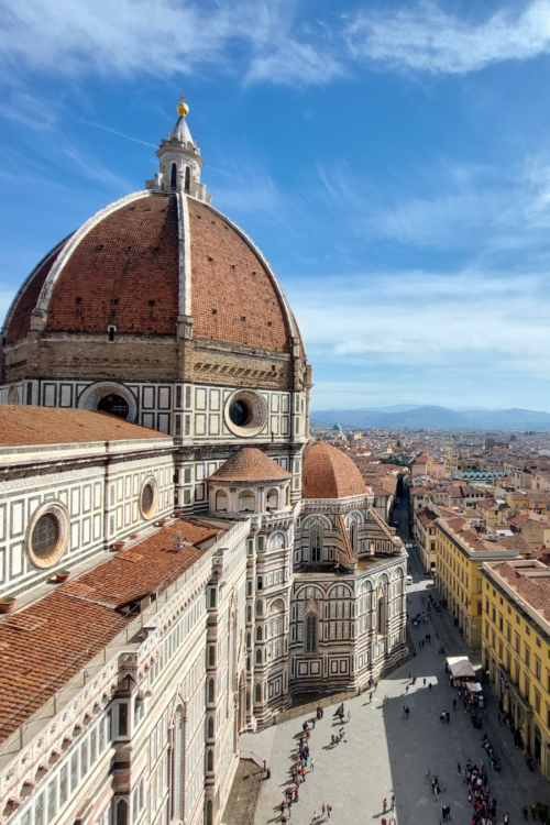 Florence skyline with Brunelleschi’s red-tiled dome and cathedral in foreground, viewed above sunlit streets and historic rooftops under blue sky.