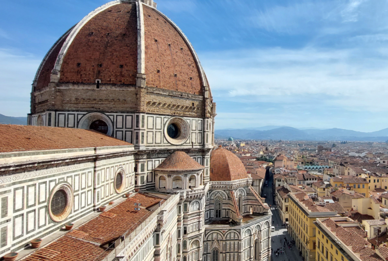 Florence skyline with Brunelleschi’s red-tiled dome and cathedral in foreground, viewed above sunlit streets and historic rooftops under blue sky.