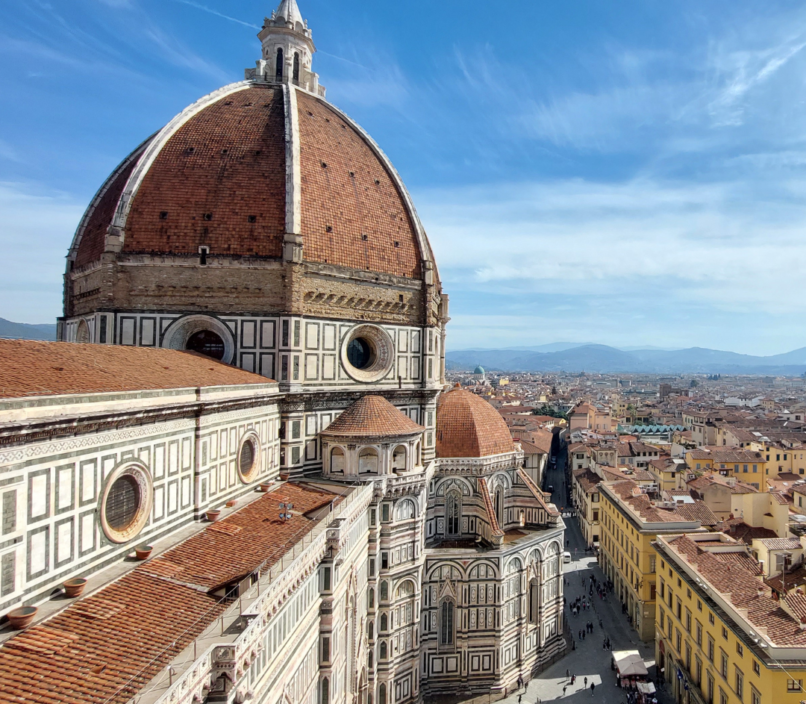 Florence skyline with Brunelleschi’s red-tiled dome and cathedral in foreground, viewed above sunlit streets and historic rooftops under blue sky.
