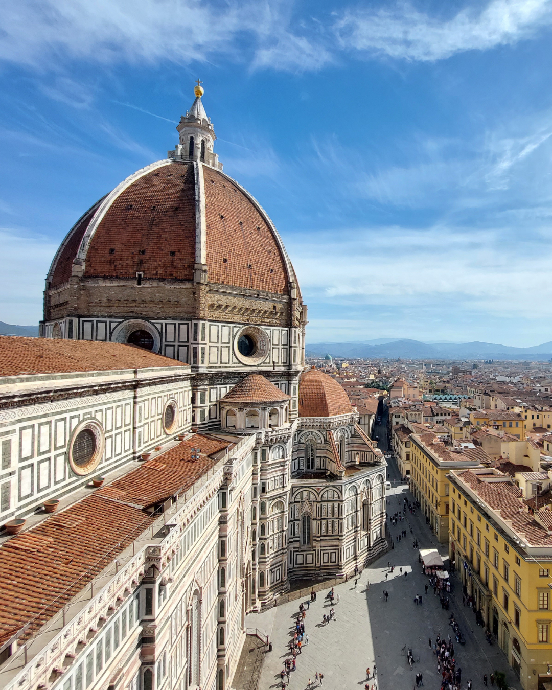 Florence skyline with Brunelleschi’s red-tiled dome and cathedral in foreground, viewed above sunlit streets and historic rooftops under blue sky.