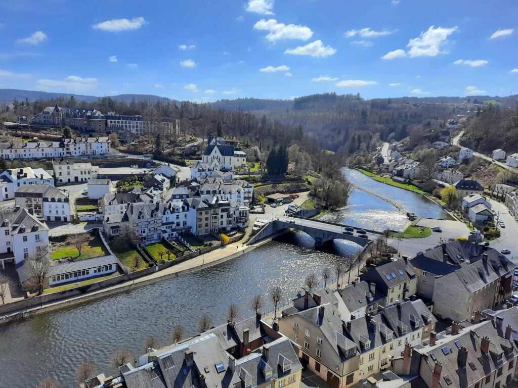 Aerial view of a historic riverside town, with slate-roofed stone houses, a low bridge, and wooded hills beneath a bright blue sky.