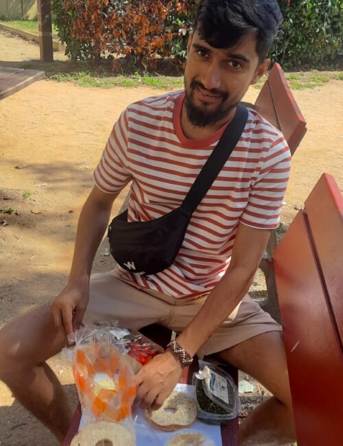 A smiling young man in a striped shirt and black crossbody bag sits on a red park bench with snacks and drinks, relaxing in a sunny garden.