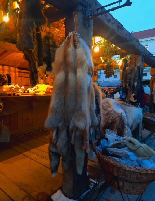 Animal pelts hang from a wooden post at a warmly lit outdoor market stall, with fur-lined baskets and shoppers creating a rustic winter-market atmosphere.