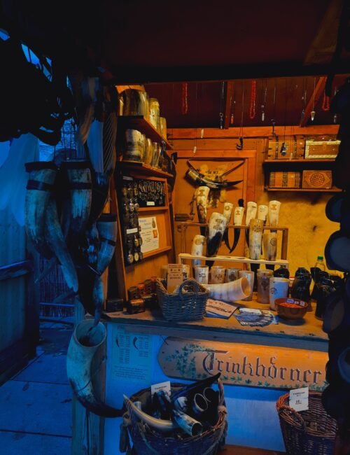 A cluttered medieval apothecary stall displays jars, bottles, herbs and tools beneath wooden shelves, glowing amber in a mysterious, blue-shadowed room.