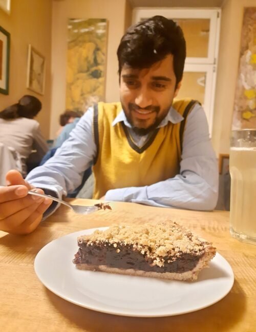 A smiling man in a yellow sweater vest sits in a cozy café, holding a fork over a slice of crumb-topped chocolate cake on a white plate.
