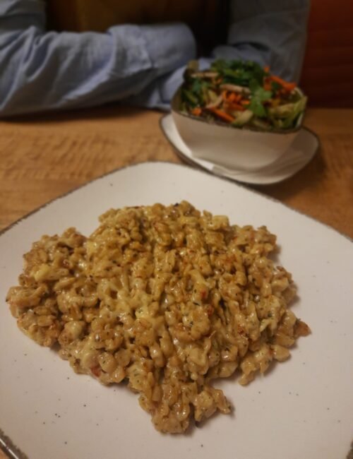 A creamy, herb-speckled pasta dish fills a white plate in the foreground, with a colorful salad behind it on a wooden table; a cozy, homemade meal.
