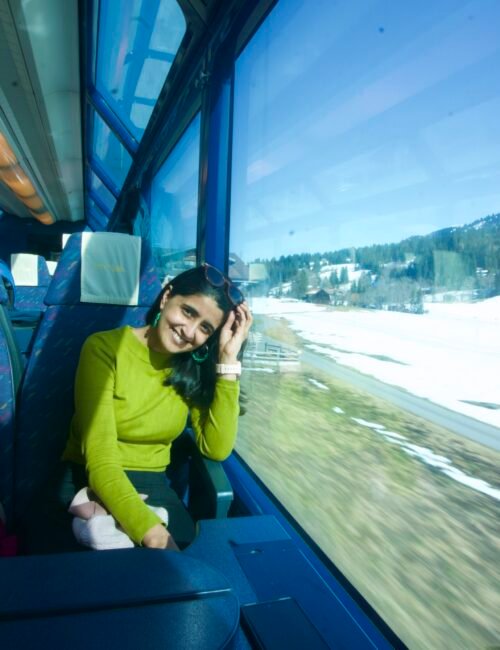 Smiling woman in a lime-green sweater sits beside a train window, leaning on her hand as snowy countryside passes in bright blue light.