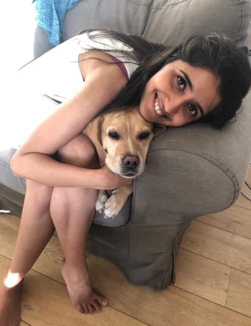 A smiling woman in a white top sits on a gray chair, cuddling a small tan dog on her lap in a cozy indoor setting.
