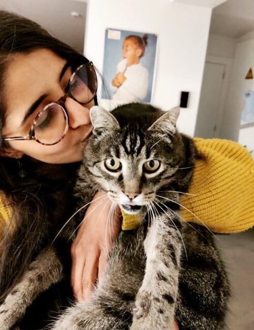 A smiling woman in glasses cuddles a tabby cat wearing a yellow collar; the cat looks at the camera in a cozy, affectionate moment.