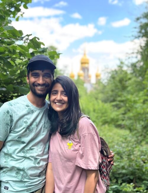 Smiling couple in casual clothes pose amid lush greenery, with a golden-domed church softly blurred behind them under a bright blue sky. The mood is warm and cheerful.