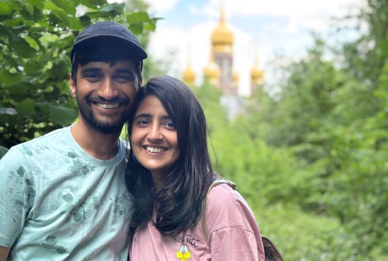 Smiling couple in casual clothes pose amid lush greenery, with a golden-domed church softly blurred behind them under a bright blue sky. The mood is warm and cheerful.