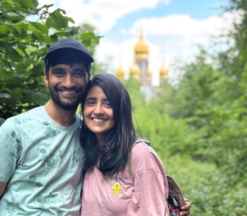 Smiling couple in casual clothes pose amid lush greenery, with a golden-domed church softly blurred behind them under a bright blue sky. The mood is warm and cheerful.