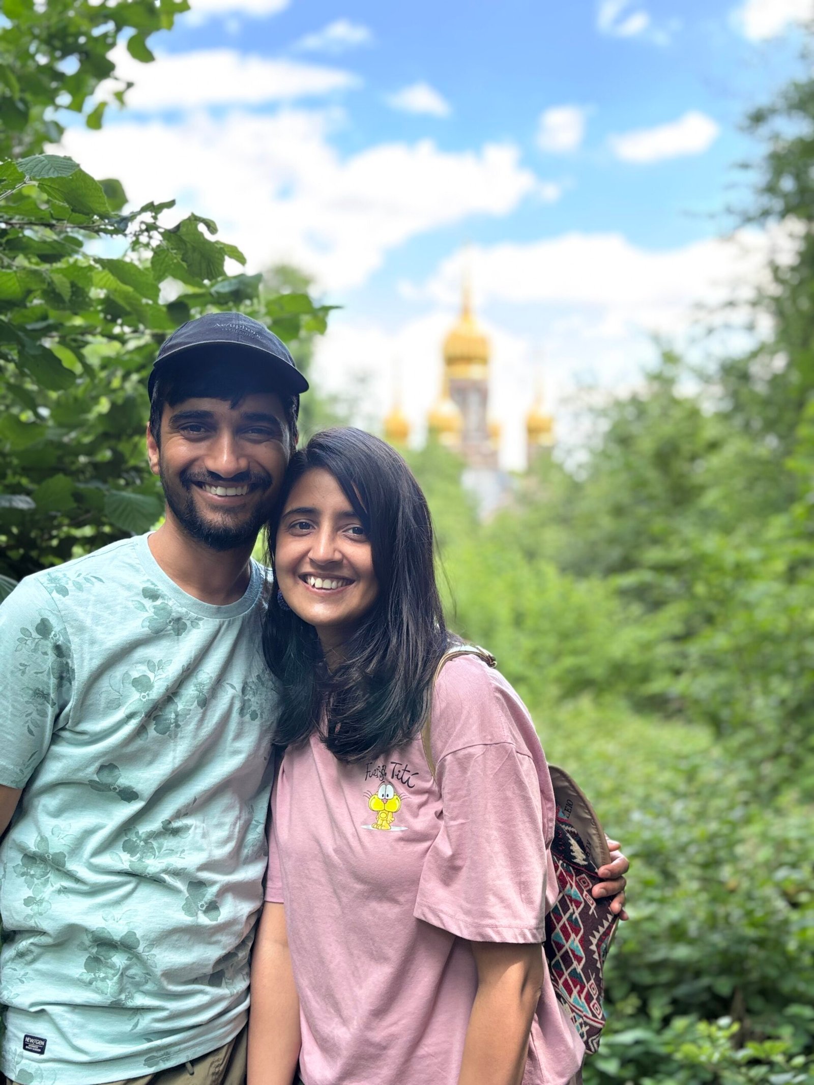 Smiling couple in casual clothes pose amid lush greenery, with a golden-domed church softly blurred behind them under a bright blue sky. The mood is warm and cheerful.