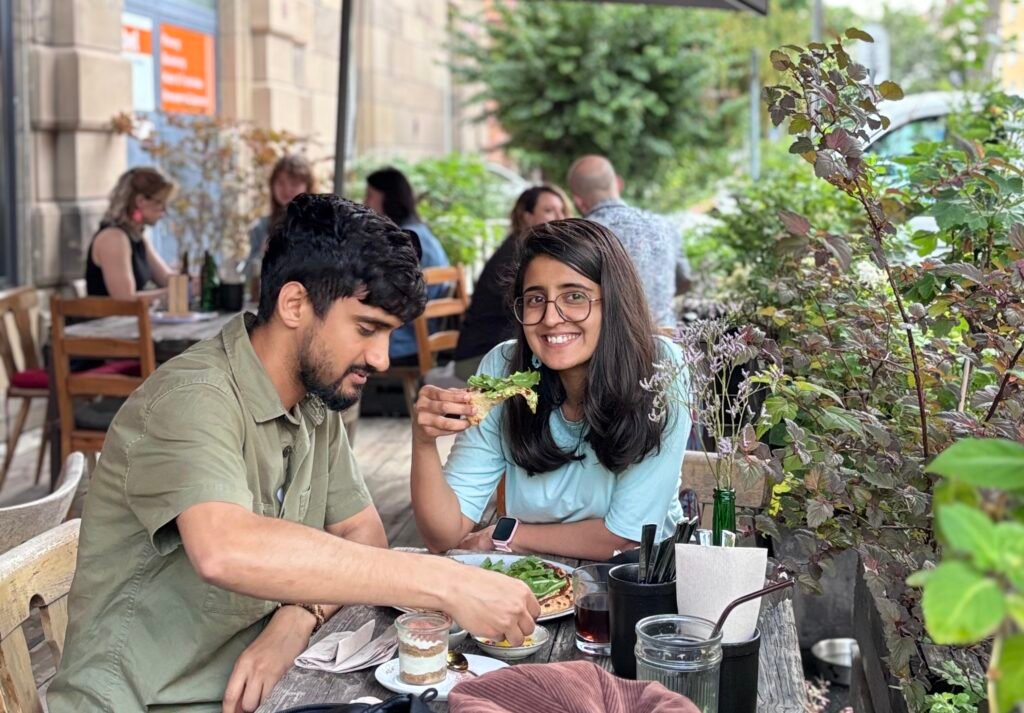 A smiling woman and man share lunch at an outdoor café, reaching for food beside leafy plants; their relaxed expressions create a warm, friendly mood.