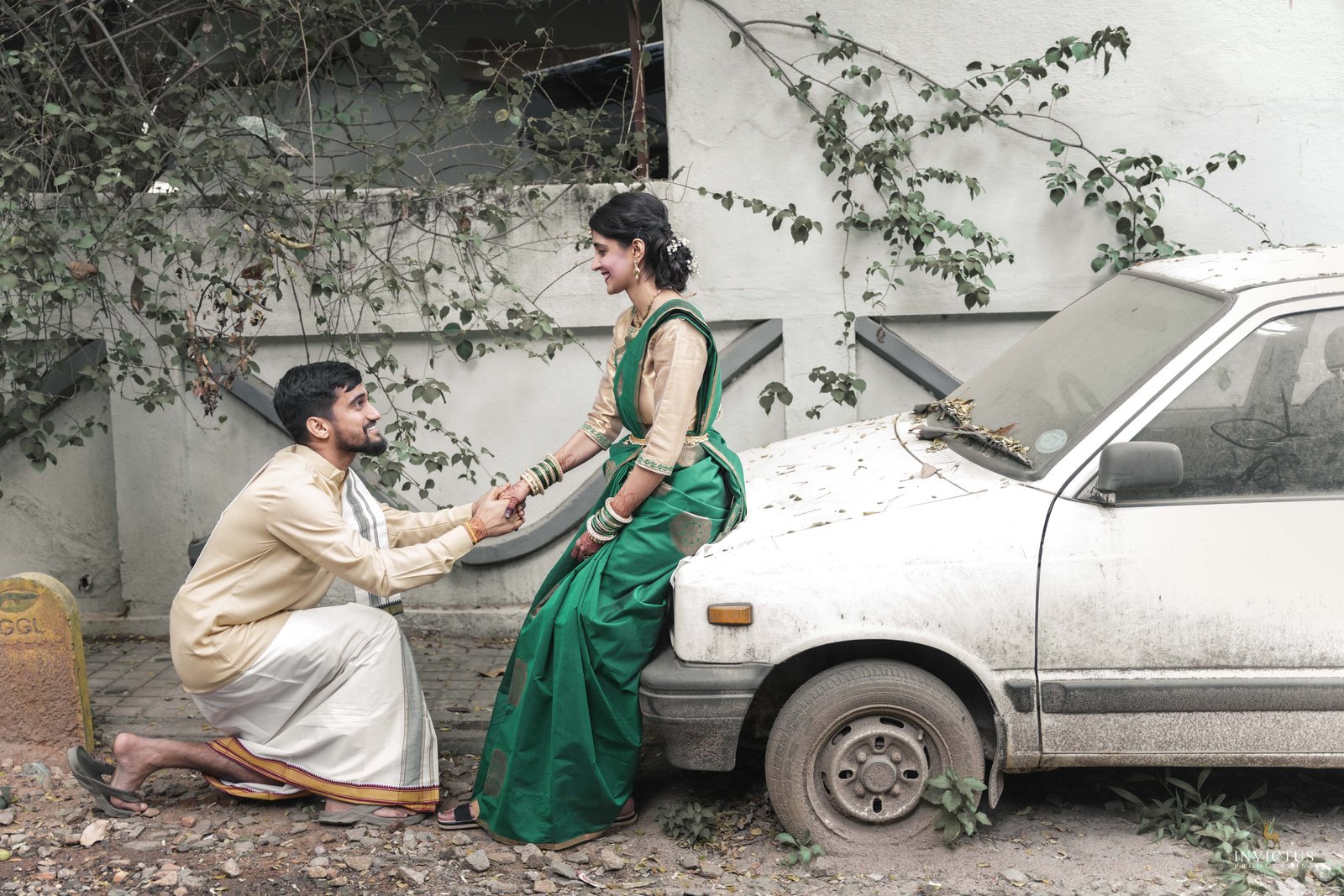 A man in cream traditional clothes kneels beside a dusty white car, holding hands with a woman in a green sari in a vine-covered courtyard; a tender scene.