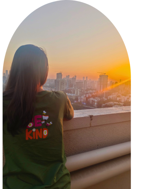 A woman in a green “Be Kind” shirt looks over a city skyline from a balcony at sunset, framed by an arch; warm light creates a calm, reflective mood.