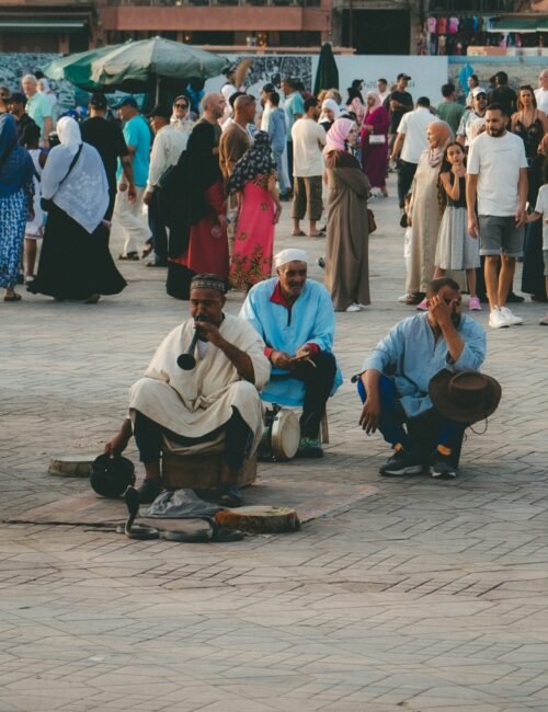Street musicians sit on a stone plaza as pedestrians pass behind them in a busy market square, creating a lively, everyday urban scene.